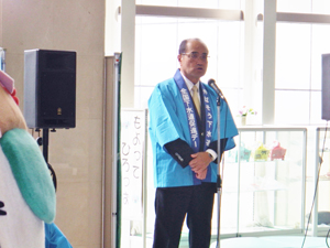 室内の中、マイクを前に青いはっぴを着た筑紫野市副市長の挨拶の写真