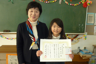賞状と副賞を両手に持って微笑む髪の長い女の子と隣に立つ女性の先生の写真